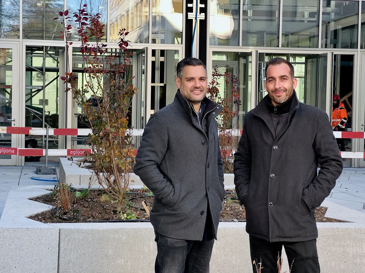 Zwei Männer mittleren Alters stehen in graue Kurzmäntel gepackt auf einem Platz vor einem Gebäude mit Fensterfronten und lächeln in die Kamera.